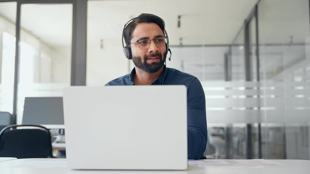 Busy Indian call center agent wearing headset talking to client working in customer support office. Professional contract service telemarketing operator using laptop having conversation.