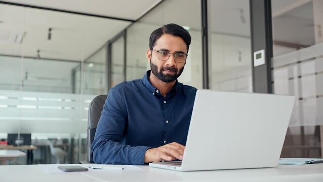 Busy smiling professional Indian business man company employee, male worker, manager or financial analyst typing on computer technology using laptop working with ai solutions sitting at office desk.