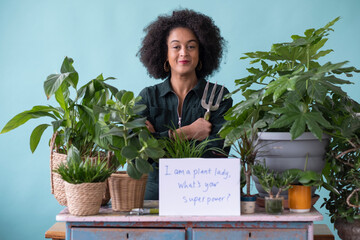 Studio portrait of smiling woman with potted plants, gardening fork and sign