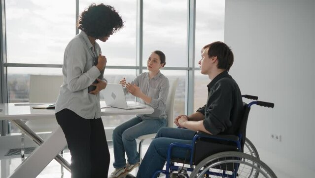 Paralyzed man works with colleagues. Guy in wheelchair has spine trauma discussing general project in office on meeting with multiethnic team women colleagues. Teamwork brainstorming, special needs.
