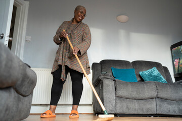 Mature woman sweeping floor in living room - obrazy, fototapety, plakaty