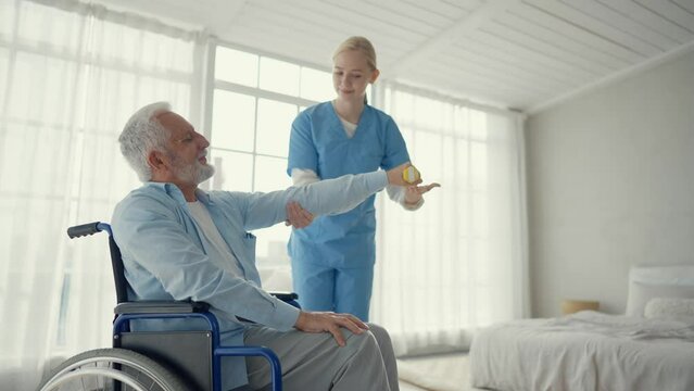 Man in wheelchair exercising with nurse. Professional medical worker woman help senior male doing physical hands exercise with dumbbell. Palliative care in late stage of neurodegenerative pathologies.