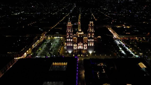 Aerial above the illuminated Morelia Cathedral with the cityscape of Morelia and the streets around