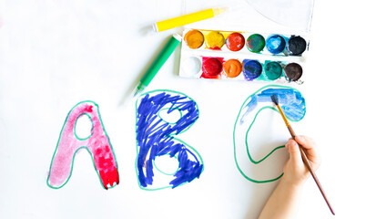 Letters of the ABC alphabet, inks, markers drawn on a white sheet of paper. A banner for school, a close-up of a child's hand, a child coloring letters on a school poster. Photo with copy space