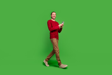 Full length photo of good mood smart guy wear red sweatshirt go empty space with smartphone in hands isolated on green color background