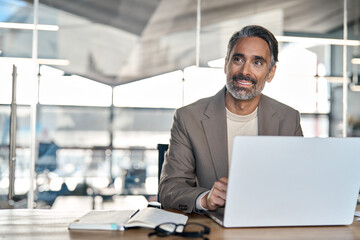 Smiling mature business man executive wearing suit sitting at desk using laptop. Happy busy professional middle aged businessman investor banker working on computer looking away in office. Copy space.