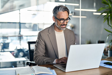 Smiling mature business man executive wearing suit sitting at desk using laptop. Happy busy professional middle aged businessman ceo investor banker working on computer corporate technology in office.