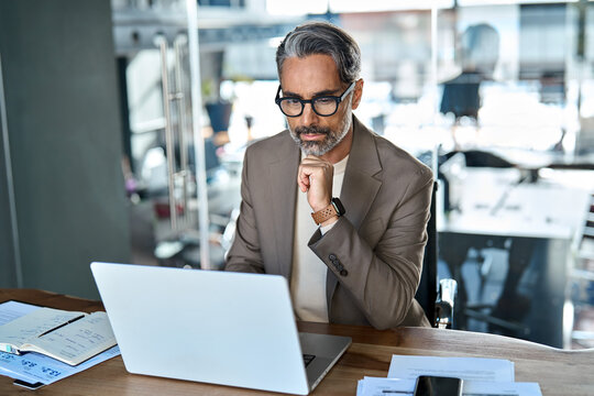 Serious busy middle aged professional business man executive sitting at desk using laptop. Busy mature businessman manager investor working on computer thinking analyzing financial data in office.