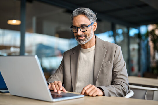 Smiling busy middle aged mature professional business man entrepreneur wearing suit and eyeglasses sitting at table outdoors working online using laptop computer hybrid working outside office.