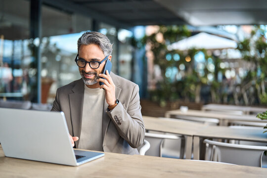 Middle aged business man talking on mobile phone hybrid working on laptop sitting outside office. Mature businessman making call on cellphone speaking to client using computer outdoors. Copy space.
