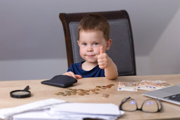 A boy in a blue t-shirt sits at a table with money, a magnifying glass and a wallet and shows a thumbs up. Business concept.