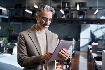 Busy mature older business man executive standing in office using digital tablet. Middle aged businessman corporate manager wearing suit working on professional financial investment project.