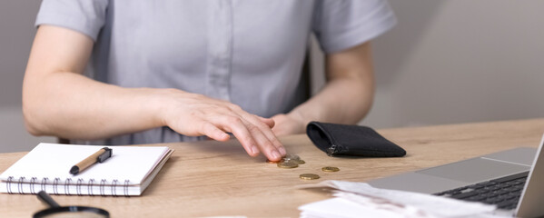 A woman counts coins poured out of her purse, cents sitting at the table. On the table is a notebook with blank sheets, a pen, a magnifying glass and a laptop. High quality photo - obrazy, fototapety, plakaty