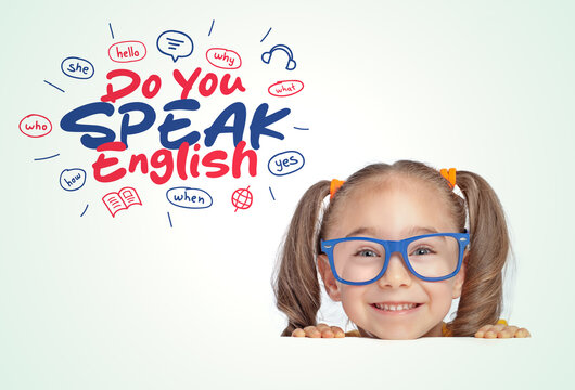 Beautiful cute little girl with eyeglasses hiding under a table, looking at the illustrations and do you speak English text above her head.  Foreign language learning concept.