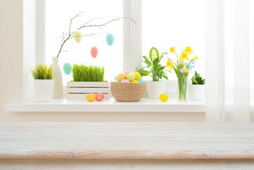 Empty easter table background in front of blurred multicolor flowers and eggs on sunny kitchen window sill