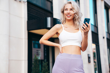 Fototapeta na wymiar Young beautiful smiling hipster woman in trendy summer clothes. Carefree model posing in street at sunset. Positive blond female. Holds smartphone, uses phone apps, looks at telephone screen