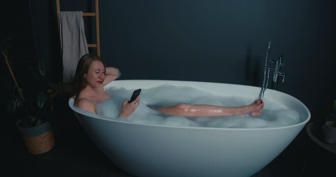 Side view happy attractive Caucasian blonde woman smiles resting in glamour bubble bath using smart phone.
