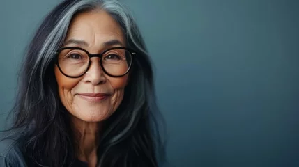 Fotobehang A smiling elderly woman with long dark hair and glasses against a soft blue background. © iuricazac