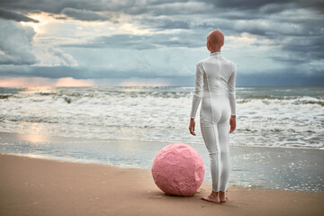 Hairless performer girl with alopecia in white futuristic suit stands on sea beach with pink sphere...