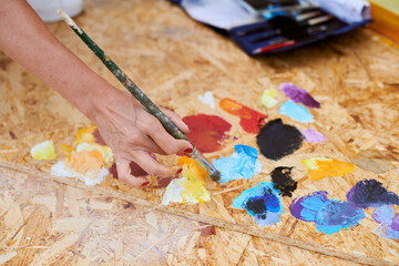 Female painter hand dips paintbrush into palette of colorful paints for live painting of picture...