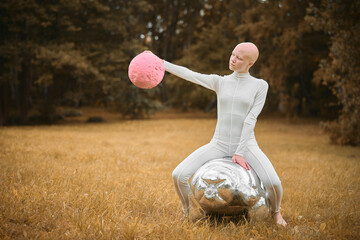 Young hairless girl with alopecia in white cloth sits on tardigrade figure and holds pink ball in...