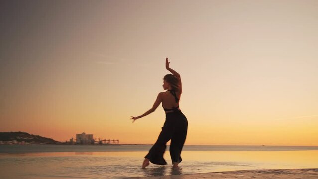 Attractive woman performing sensual dance in water. Young female dancer in black dress rehearsing under sunset sky. Lady dancing gracefully in nature.