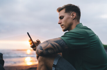 Thoughtful man with smartphone sitting at seashore in sunset