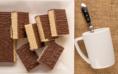 Several sweet chocolate wafers with ceramic saucer and cup on jute cloth, macro, top view.