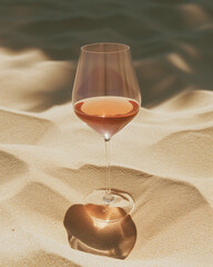 Rose wine in an elegant glass is placed on the sand. The sunlight passes through the glass and reflects the light onto the sand.