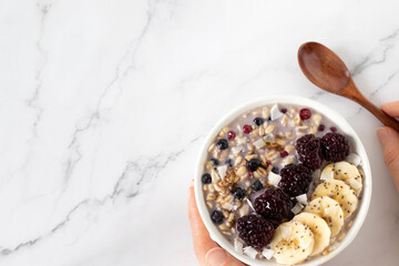 Hand holding overnight oatmeal bowl with berries, banana, chia seeds, and granola with wooden spoon on white kitchen table. Top view. Copy space. Eating healthy plant-based vegan breakfast meal.