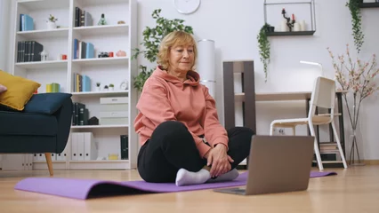 Fotobehang A senior woman communicates with a fitness trainer online, exercising at home © Synthex🇺🇦