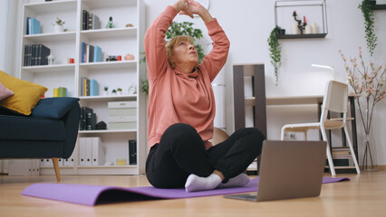A mature woman engages in fitness training online, sitting on a mat and exercising at home