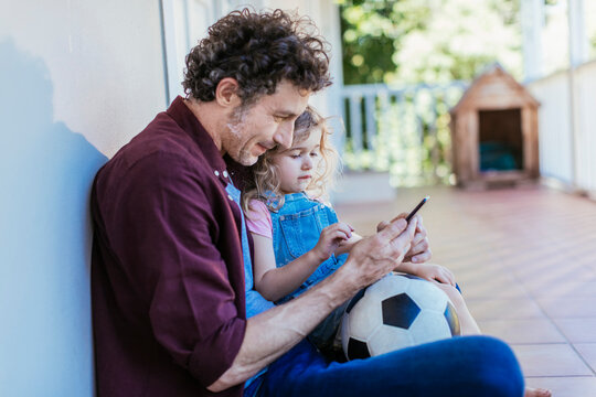 Father and daughter with soccer ball looking at smartphone outdoors - Powered by Adobe