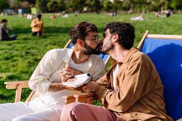 Loving gay couple sitting on deckchair and kissing in park