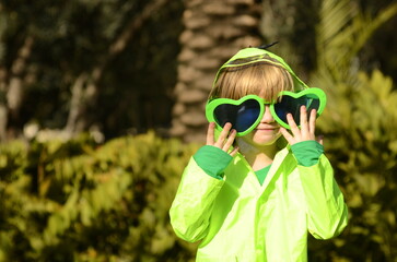 Cute boy, 5 years old, wearing big green heart-shaped glasses. Funny face. Palm trees and sky in...