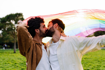 Happy gay couple kissing and holding LGBT rainbow flag while standing in park