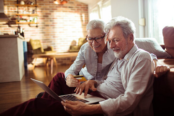 Happy senior couple using credit card on laptop at home - obrazy, fototapety, plakaty