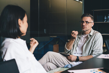 Caucasian man in optical eyeglasses for vision protection holding digital tablet and have conversation with colleague, male employee in eyewear and earbuds discussing business ideas with partner
