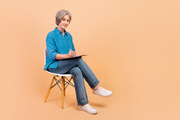 Full length photo of focused person wear stylish shirt writing checklist sit on chair on interview isolated on beige color background