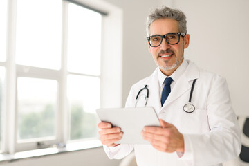 Senior doctor with glasses and a friendly demeanor holds a tablet, standing by a window with natural light that suggests a modern and approachable healthcare setting. Contemporary medical practice