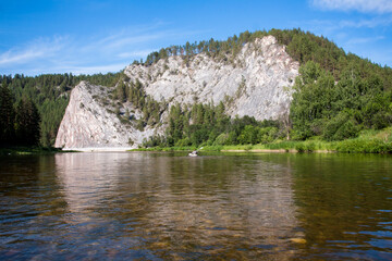 Fototapeta na wymiar Tranquil River Scene with Rocky Cliff, Lush Green Trees, and Serene Blue Sky
