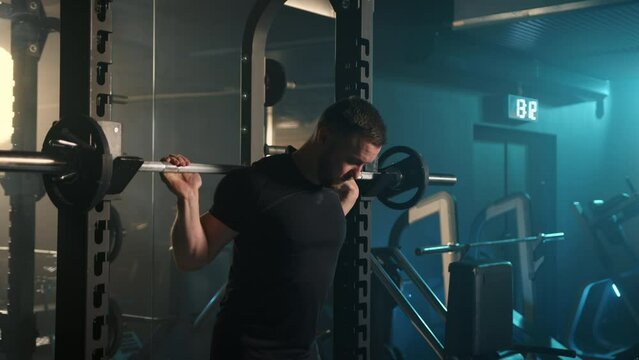 In the mood-lit gym, a man commits to his weightlifting regimen, focusing on barbell squats that challenge his lower body strength, all under the watchful glow of blue and orange lights.