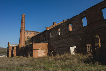 Fototapeta na wymiar Ruins of an old factory building with tall chimney and overgrown vegetation under clear blue sky