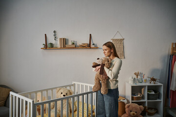helpless and upset woman with soft toy standing near crib with in bleak nursery room at home