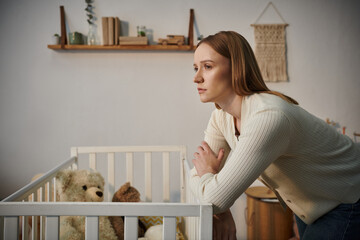 disheartened woman standing near crib with soft toys in dark nursery room at home, grieving