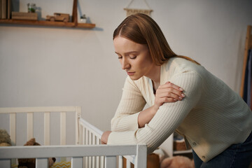 depressed woman standing near crib with soft toys in bleak nursery room at home, frustration