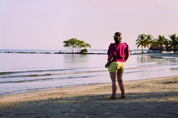 middle-aged woman dressed in yellow shorts and purple blouse standing on the beach facing the sea with her back to us on the beach facing the sea and looking into the distance.
