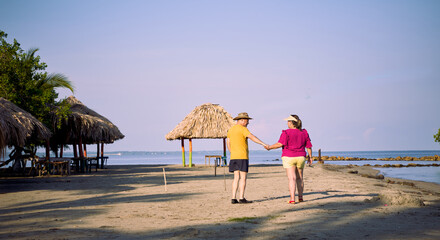 An elderly couple holding hands, dressed in beach attire; he wears a turned hat and she wears a large visor. They walk along the beach surrounded by thatched roofs and coastal trees, facing the sea