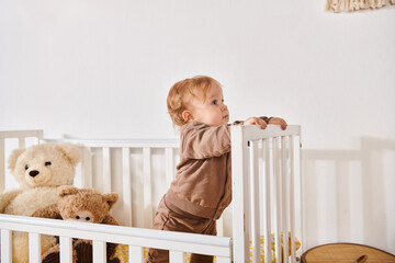 curious baby boy standing in crib with soft toys in cozy nursery room at home, blissful childhood