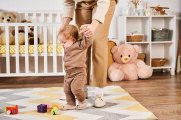 mother holding hands of toddler son walking near toys and crib in nursery room, care and support